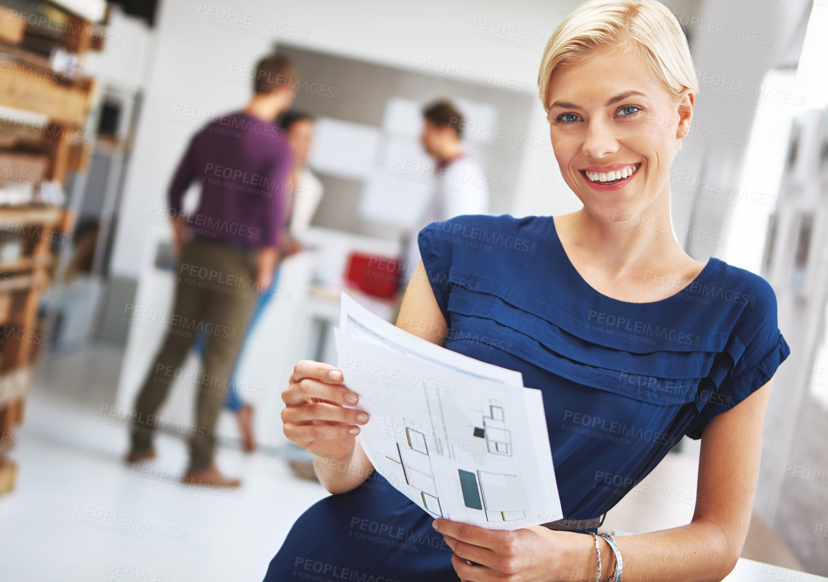 Buy stock photo Cropped portrait of a young female designer looking over some paperwork with her colleagues in the background