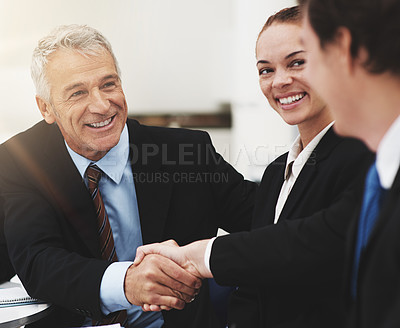 Buy stock photo Shot of two businessman shaking hands at a boardroom table while a colleague looks on
