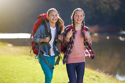 Buy stock photo Shot of two young girls wearing backpacks walking together beside a lake