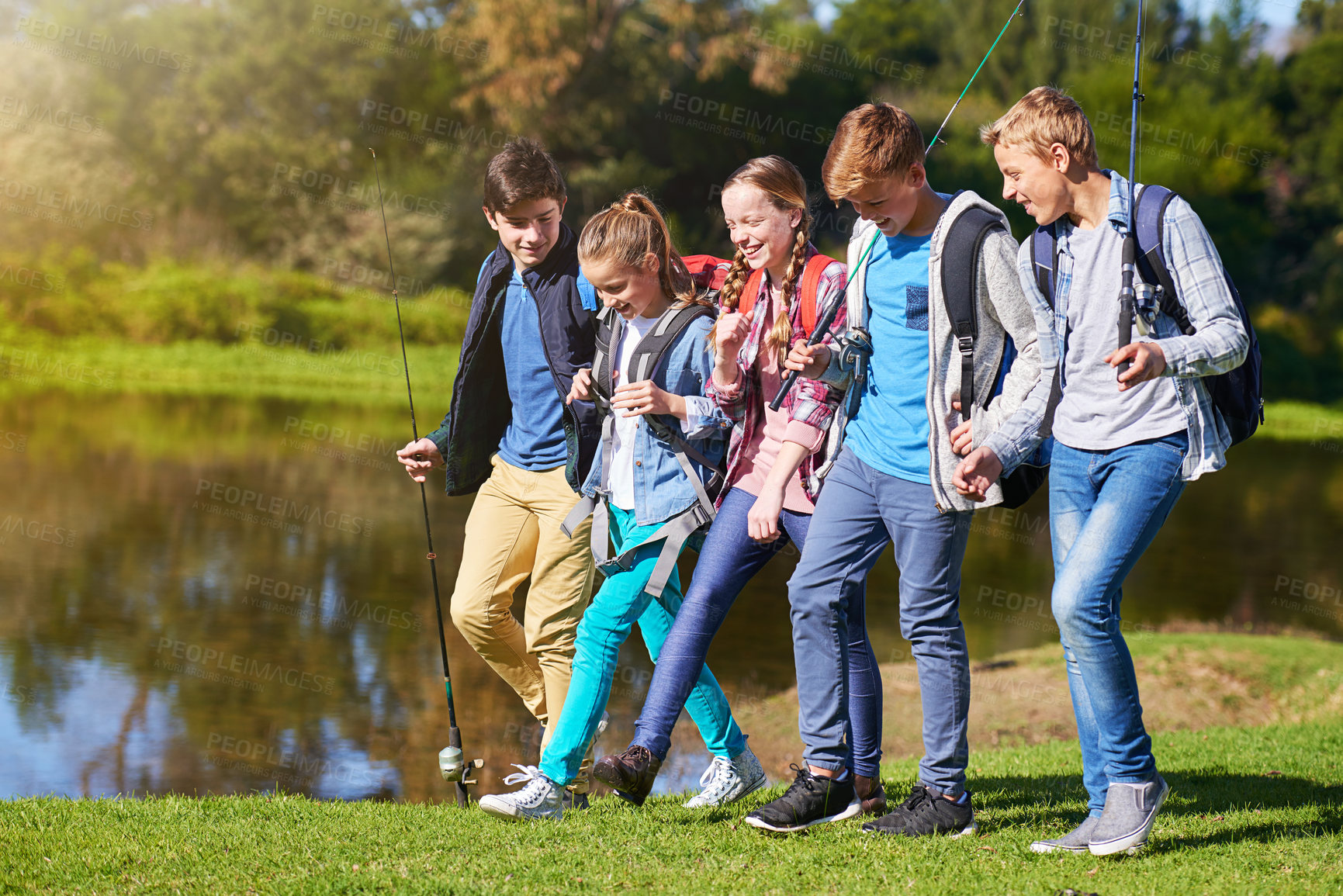 Buy stock photo A group of young friends wearing backpacks walking together beside a lake