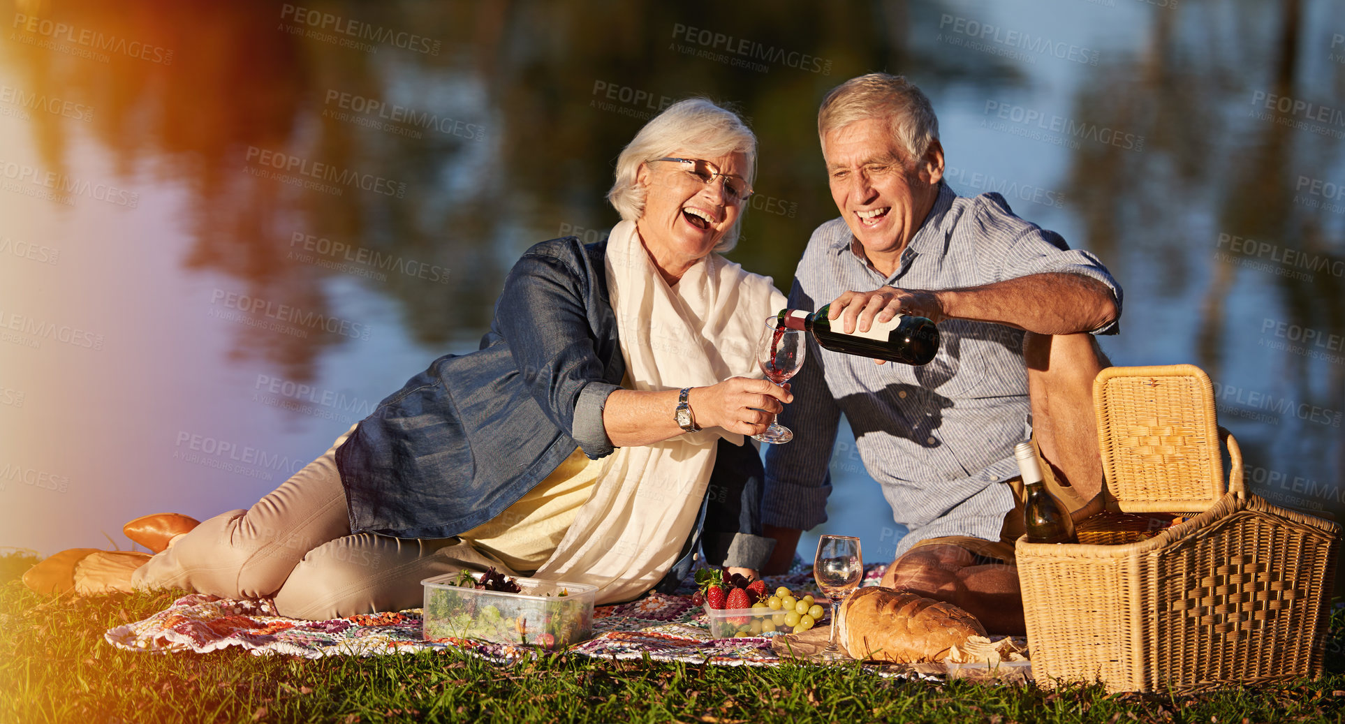 Buy stock photo Senior couple, happy and pouring wine for picnic date, sundowners or lunch by the lake. Elderly man, smiling and mature woman laughing for retirement, relationship celebration or evening drinking