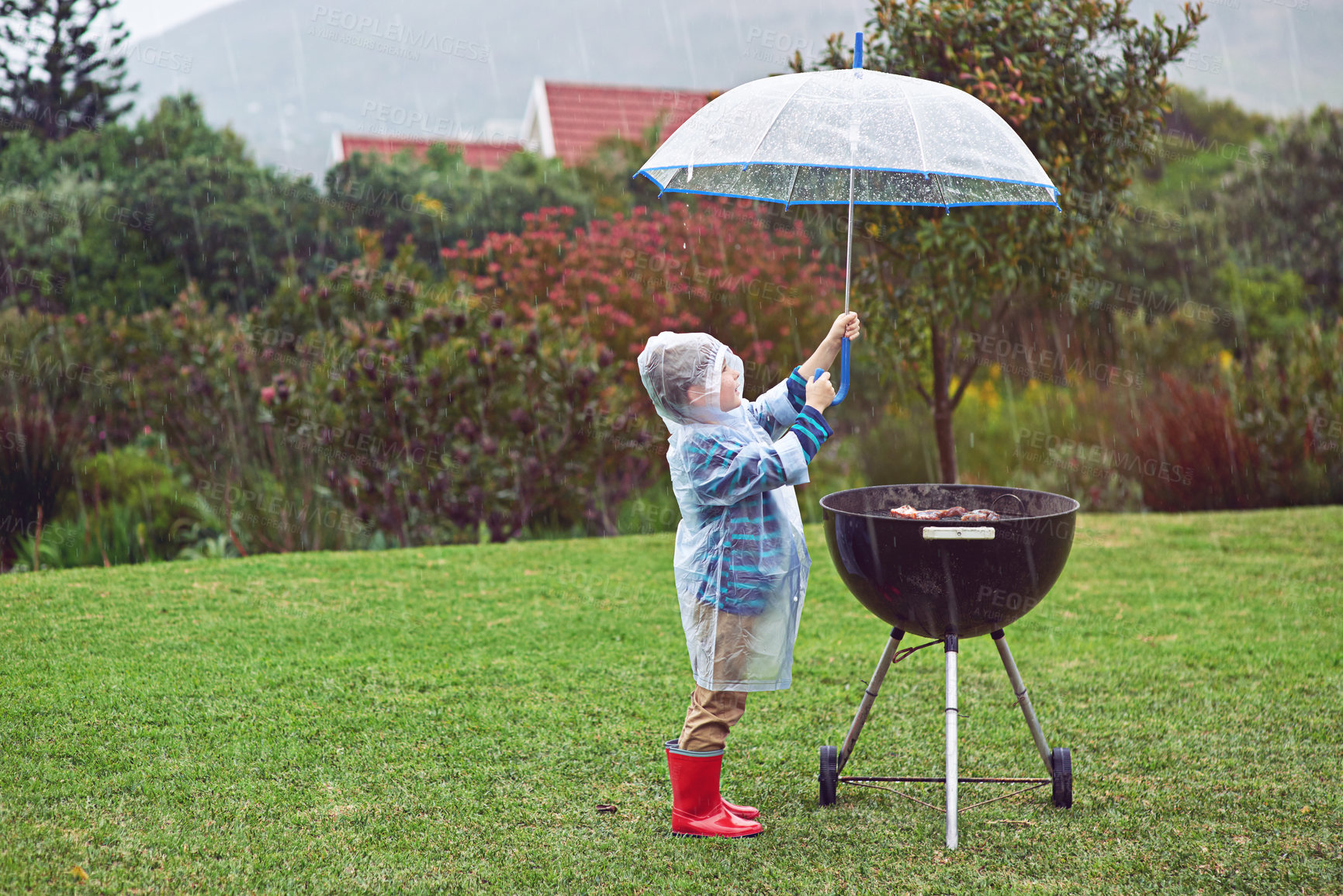 Buy stock photo Full length shot of a young boy standing in front of an outdoor grill in the rain