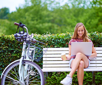 Buy stock photo Shot of a young woman using a laptop on a bench while out for a cycle in the park