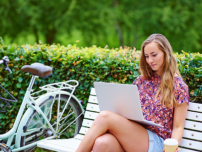 Buy stock photo Shot of a young woman using a laptop on a bench while out for a cycle in the park