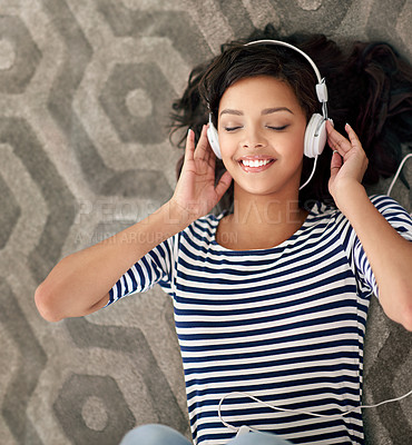 Buy stock photo High angle shot of a young woman listening to music on her cellphone while lying on the floor
