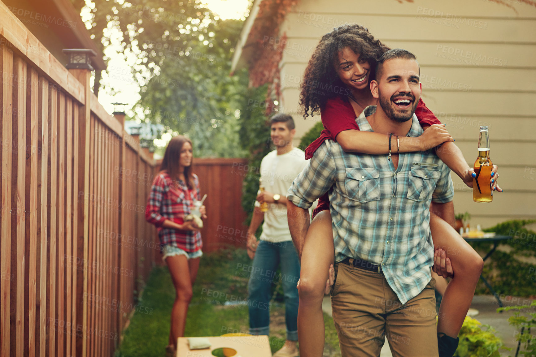 Buy stock photo Shot of a young man piggybacking his girlfriend with people standing in the background