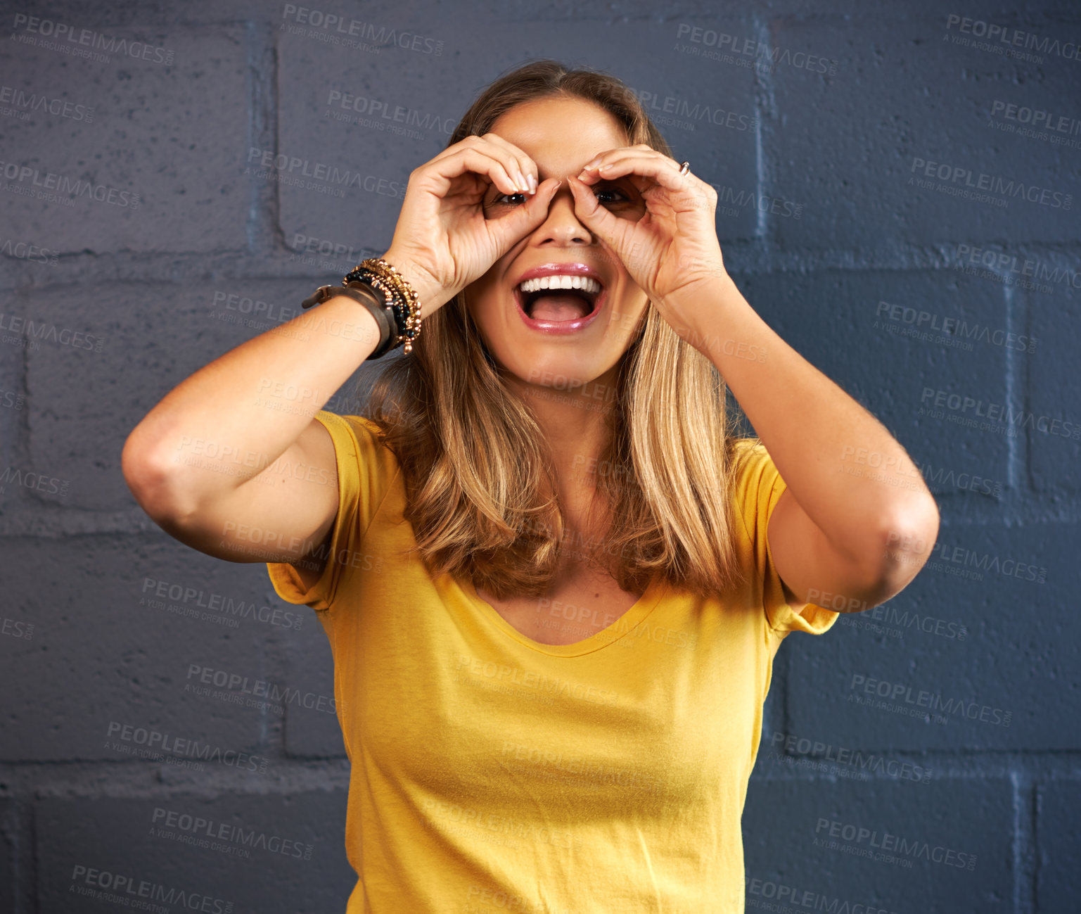 Buy stock photo Woman, portrait smile and binocular hands for fun with eyes, wellness and comedy by brick wall. Female person, happiness and playful with funny gesture, excited and isolated by dark background