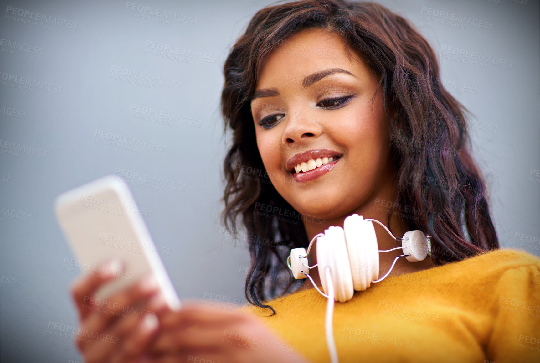Buy stock photo Studio shot of a young woman with headphones around her neck using a mobile phone against a gray background