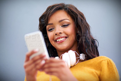 Buy stock photo Studio shot of a young woman with headphones around her neck using a mobile phone against a gray background
