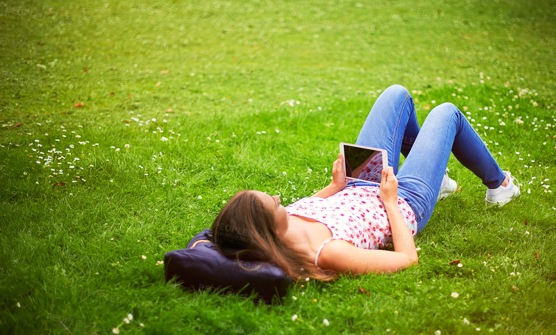 Buy stock photo Shot of a young woman using her digital tablet while lying on the grass at the park