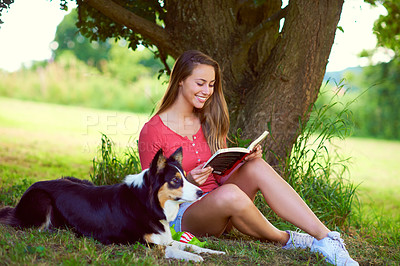 Buy stock photo Shot of a young woman reading a book while sitting with her dog under a tree