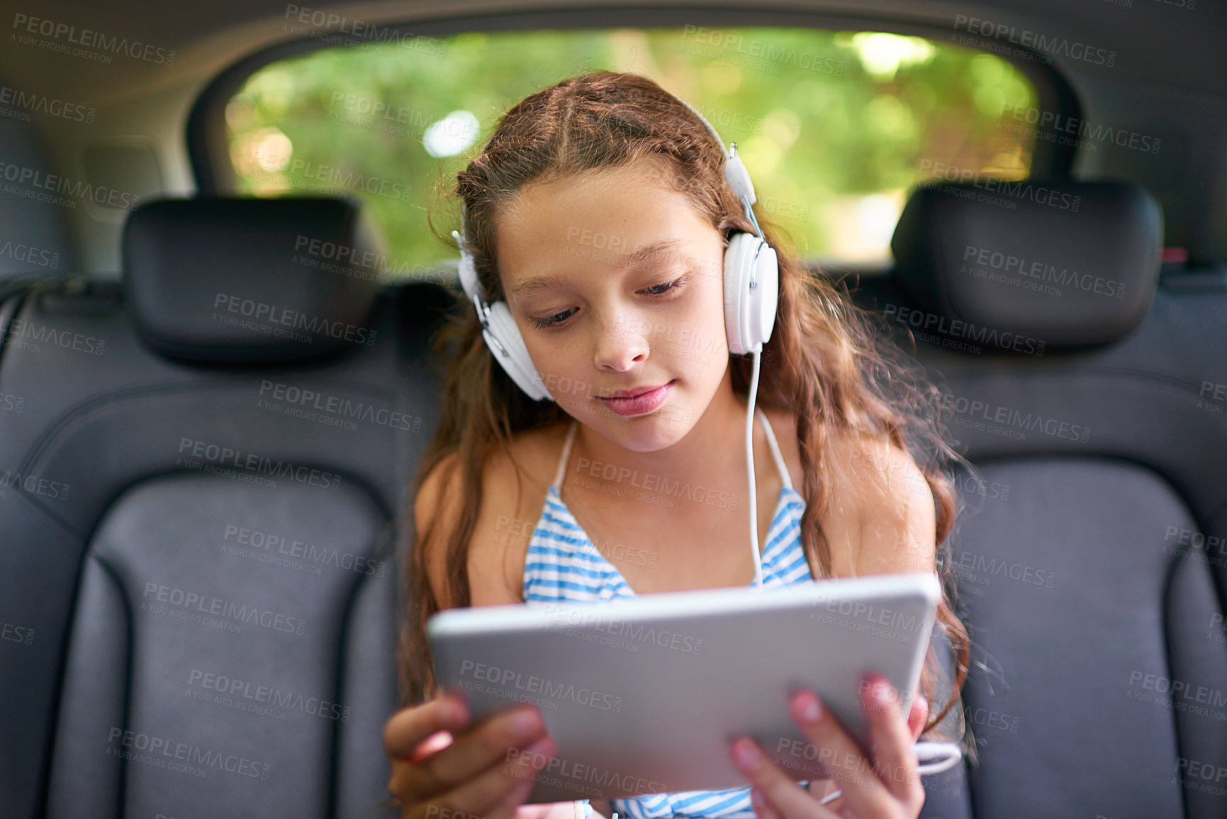 Buy stock photo Shot of a young girl sitting in a car backseat wearing headphones and using a digital tablet