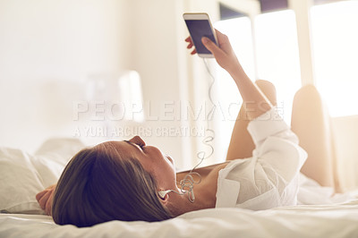 Buy stock photo Shot of a young woman listening to music on her phone while relaxing on her bed