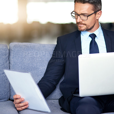Buy stock photo Shot of a businessman reading a document and using a laptop on the sofa