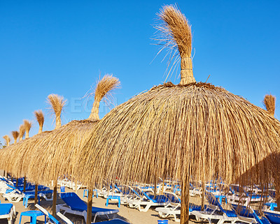 Buy stock photo Straw umbrella, chairs and blue sky at beach for travel, shade or service at luxury resort. Thatch sunshade, seats or sunshine at ocean for holiday, vacation or eco friendly design for relax in Spain