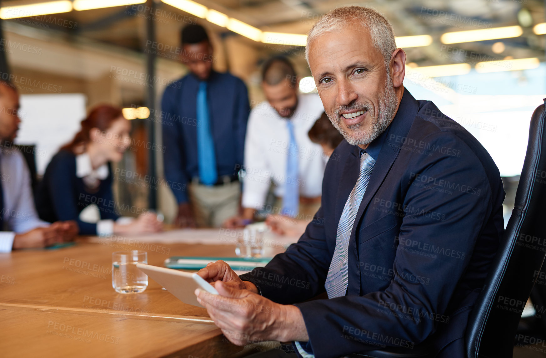 Buy stock photo Shot of a mature businessman sitting with a tablet with his  colleagues working in the background