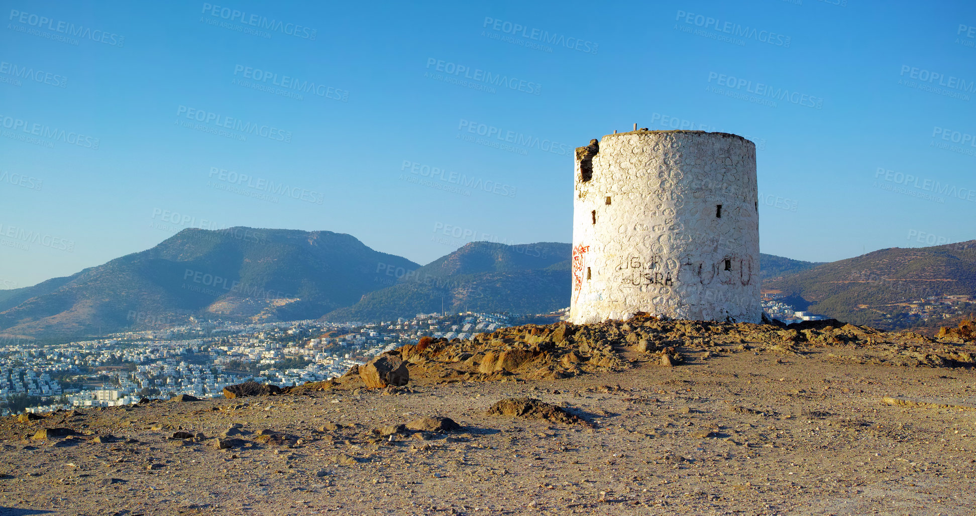 Buy stock photo Ruins of broken windmill on a hill overlooking a city surrounded by mountains. Crumbling walls of abandoned lighthouse tower. A weathered round white stone building. Architecture of an old structure