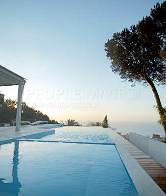 Buy stock photo An infinity swimming pool with an ocean view and blue sky at sunset. Luxury outdoor pool with trees and plants on a patio or rooftop of a holiday home or hotel. A modern pool overlooking the horizon