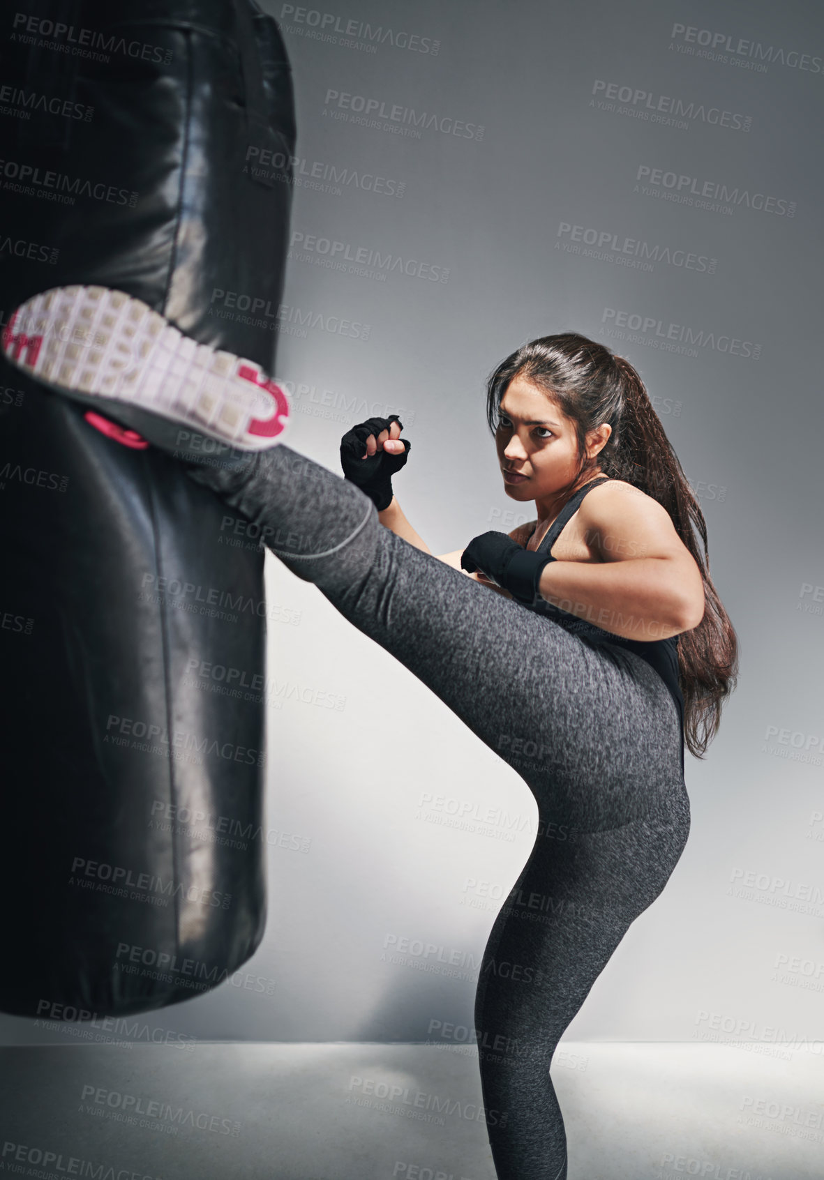 Buy stock photo Girl, kick and punching bag in studio for training, cardio boxing and building endurance for strength. Woman, serious and gray background for self defense, adrenaline control and combat workout.