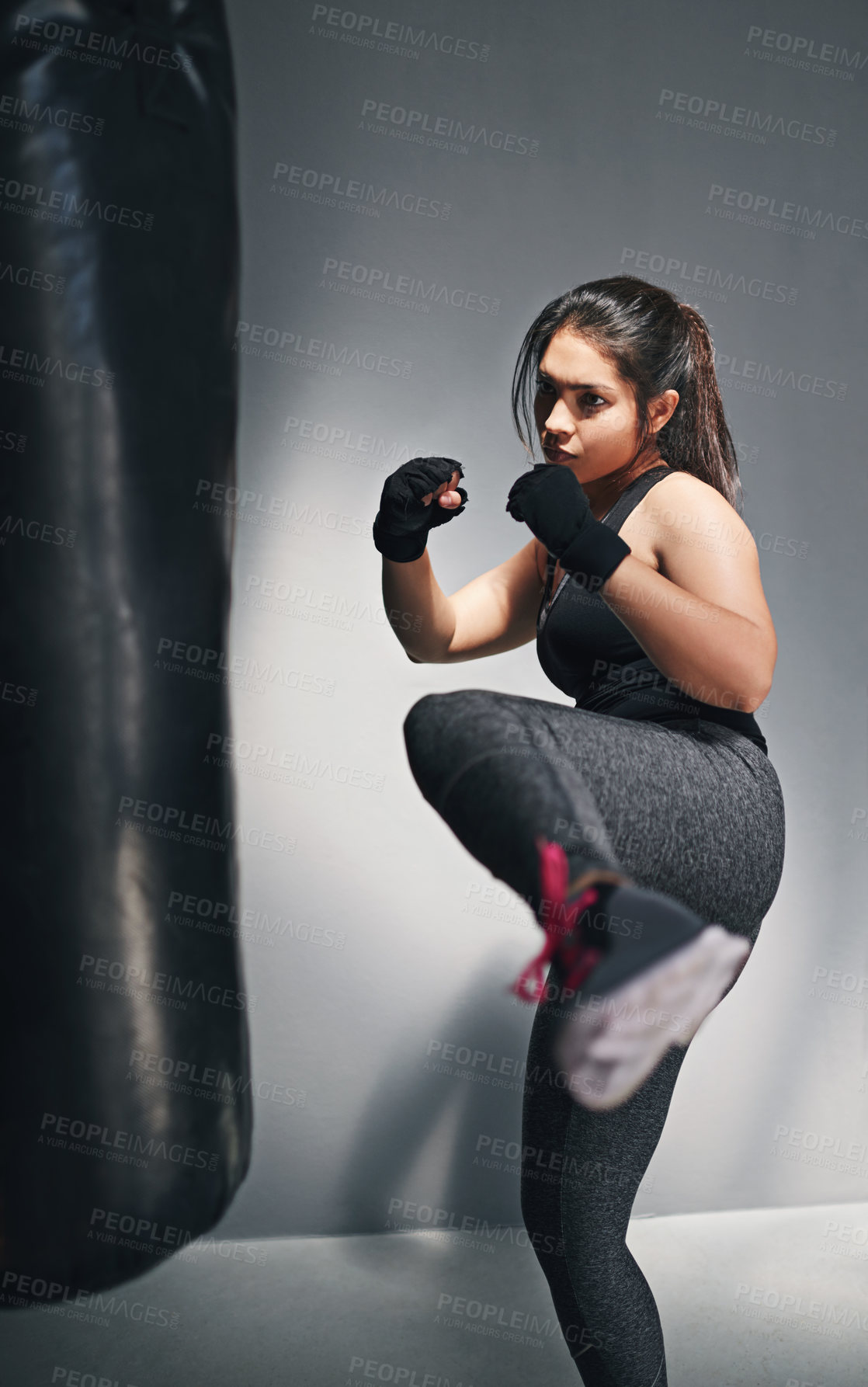 Buy stock photo Woman, kick and punching bag in studio for training, cardio boxing and building endurance for strength. Girl, serious and gray background for self defense, adrenaline control and combat workout.