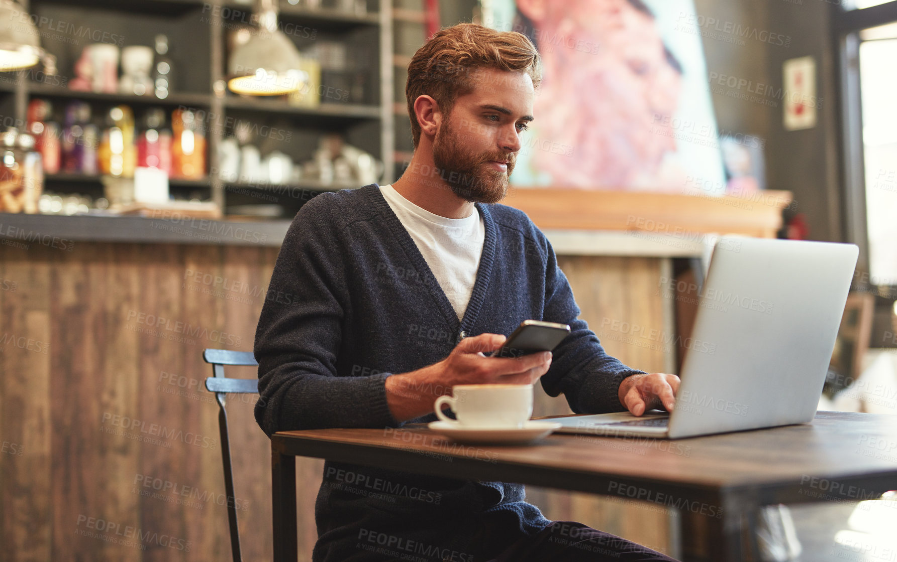 Buy stock photo Shot of a young man using a laptop and phone in a cafe