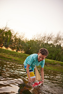 Buy stock photo Shot of a young boy standing in a lake playing with a toy boat