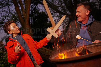 Buy stock photo Shot of a father and son playing with wood swords while toasting marshmallows over a fire