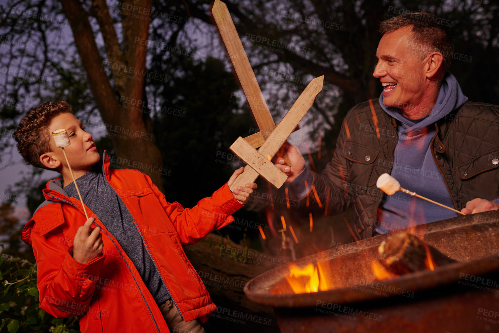 Buy stock photo Shot of a father and son playing with wood swords while toasting marshmallows over a fire