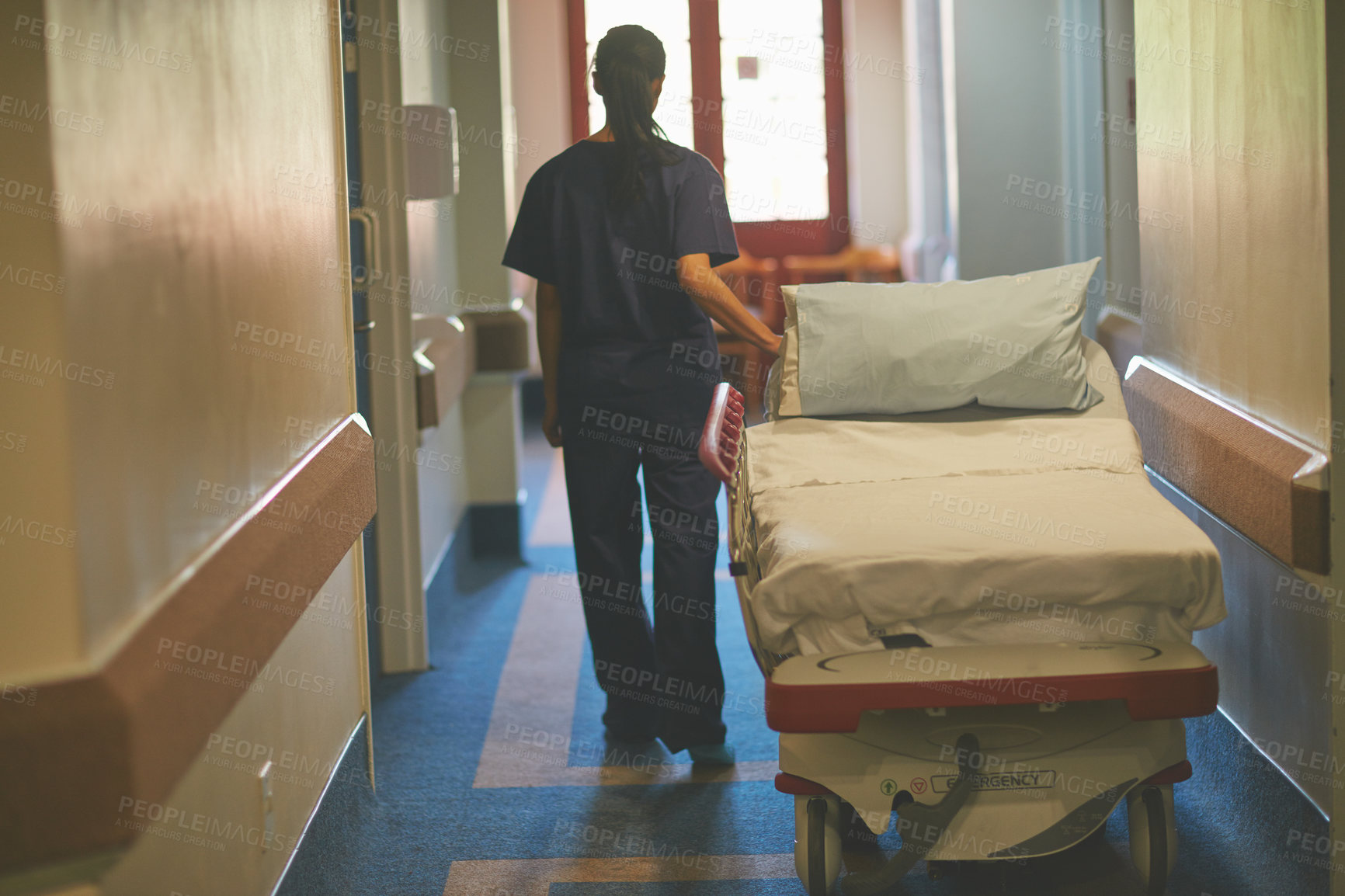 Buy stock photo Rearview shot of a female nurse pushing a hospital bed down the corridor