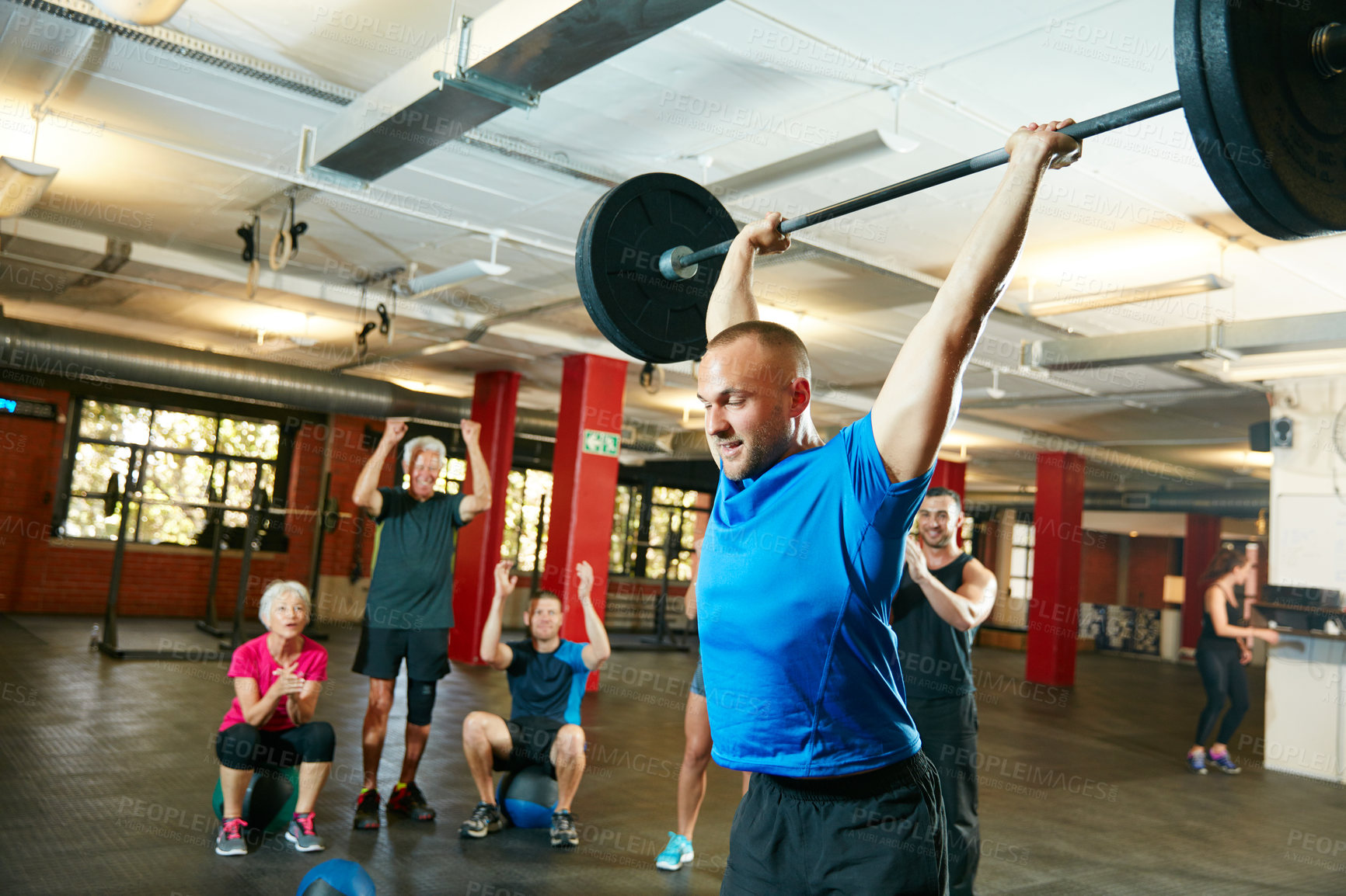 Buy stock photo Shot of a young man lifting weights while a group of people in the background watch on