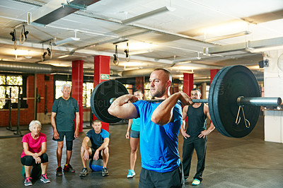 Buy stock photo Shot of a young man lifting weights while a group of people in the background watch on