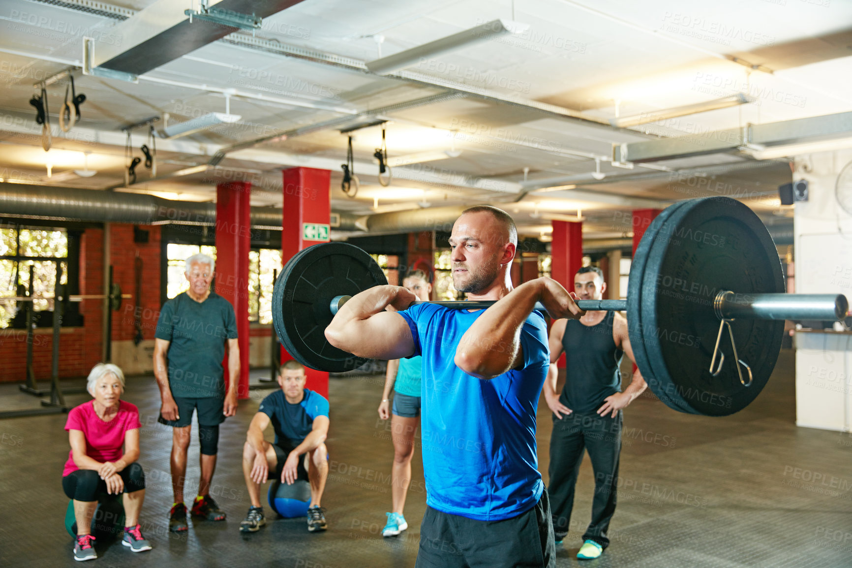 Buy stock photo Shot of a young man lifting weights while a group of people in the background watch on