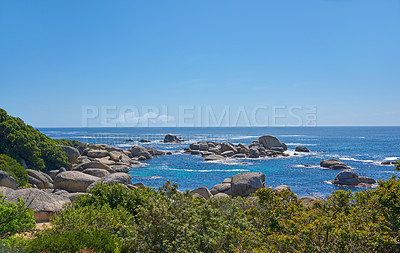 Buy stock photo Landscape of rocks in the ocean on a hot summer day in Hout Bay, Cape Town. Calm waves splashing against rocky and shrubby coastline. Beautiful large shoreline rocks by the sea with lush vegetation