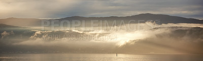 Buy stock photo Sunrise breaking through thick clouds over a calm body of water. Still, tranquil ocean in early morning. Concept of calm before the storm, rays peaking through clearing clouds with sky copyspace