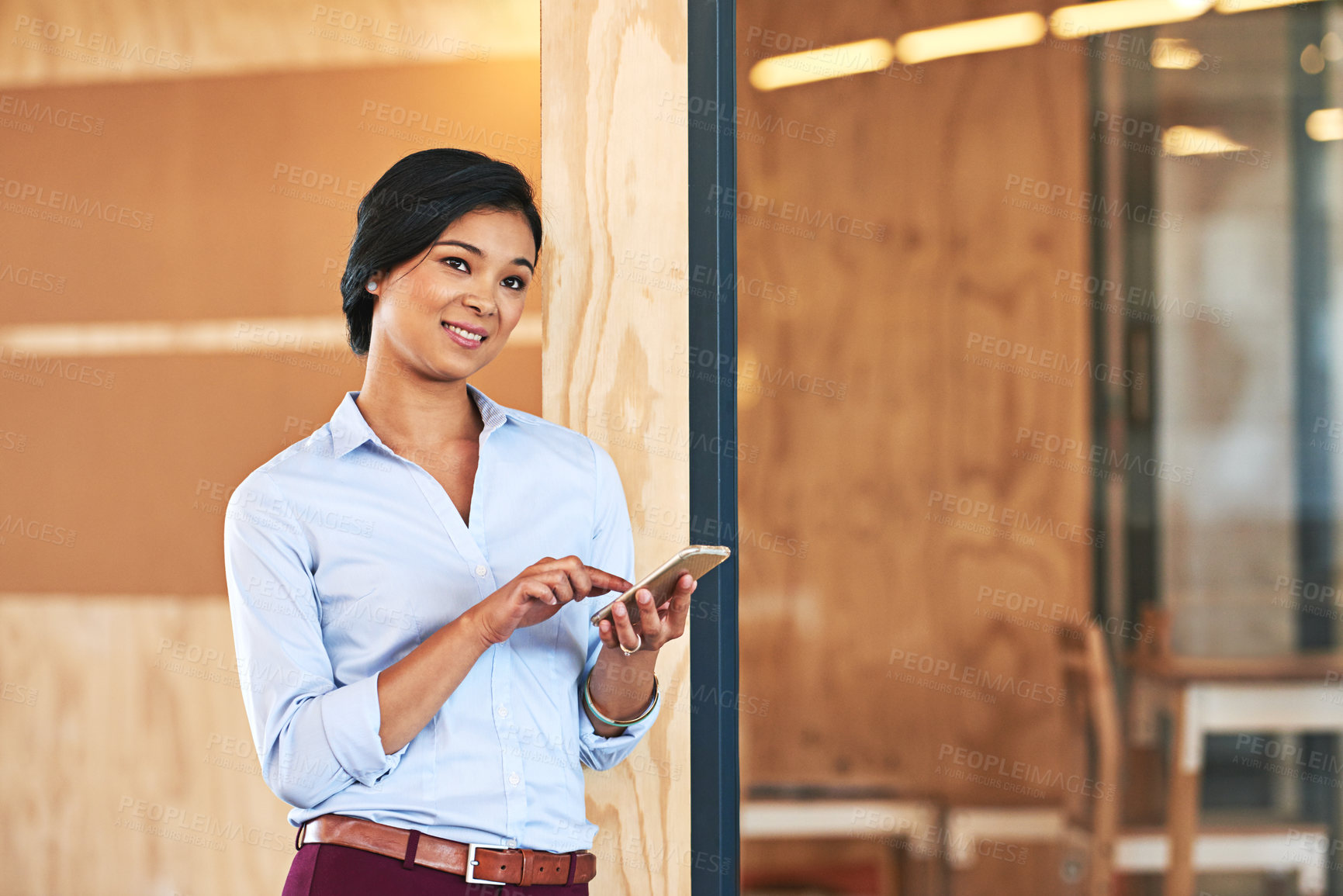 Buy stock photo Shot of a smiling young woman using her cellphone while standing in an office