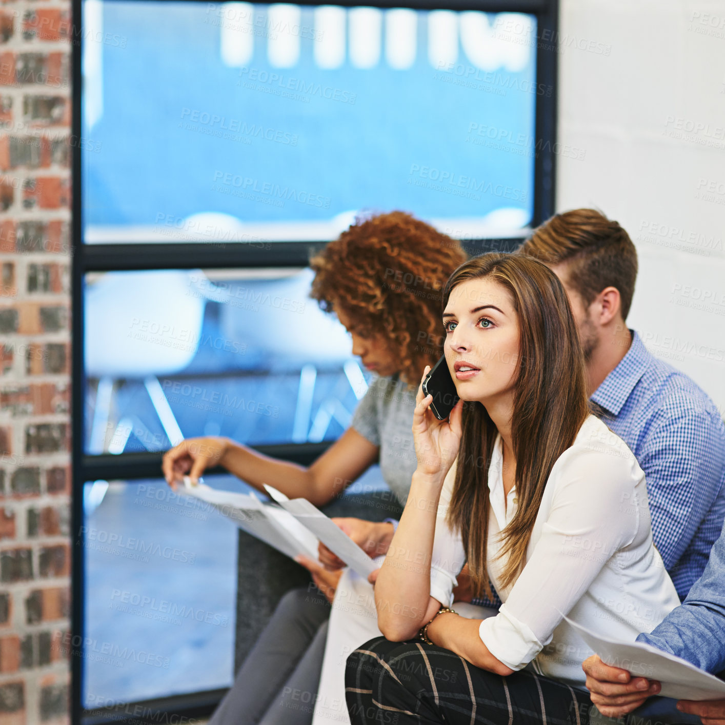 Buy stock photo Shot of a young woman talking on her phone wile sitting on a sofa with coworkers in an office