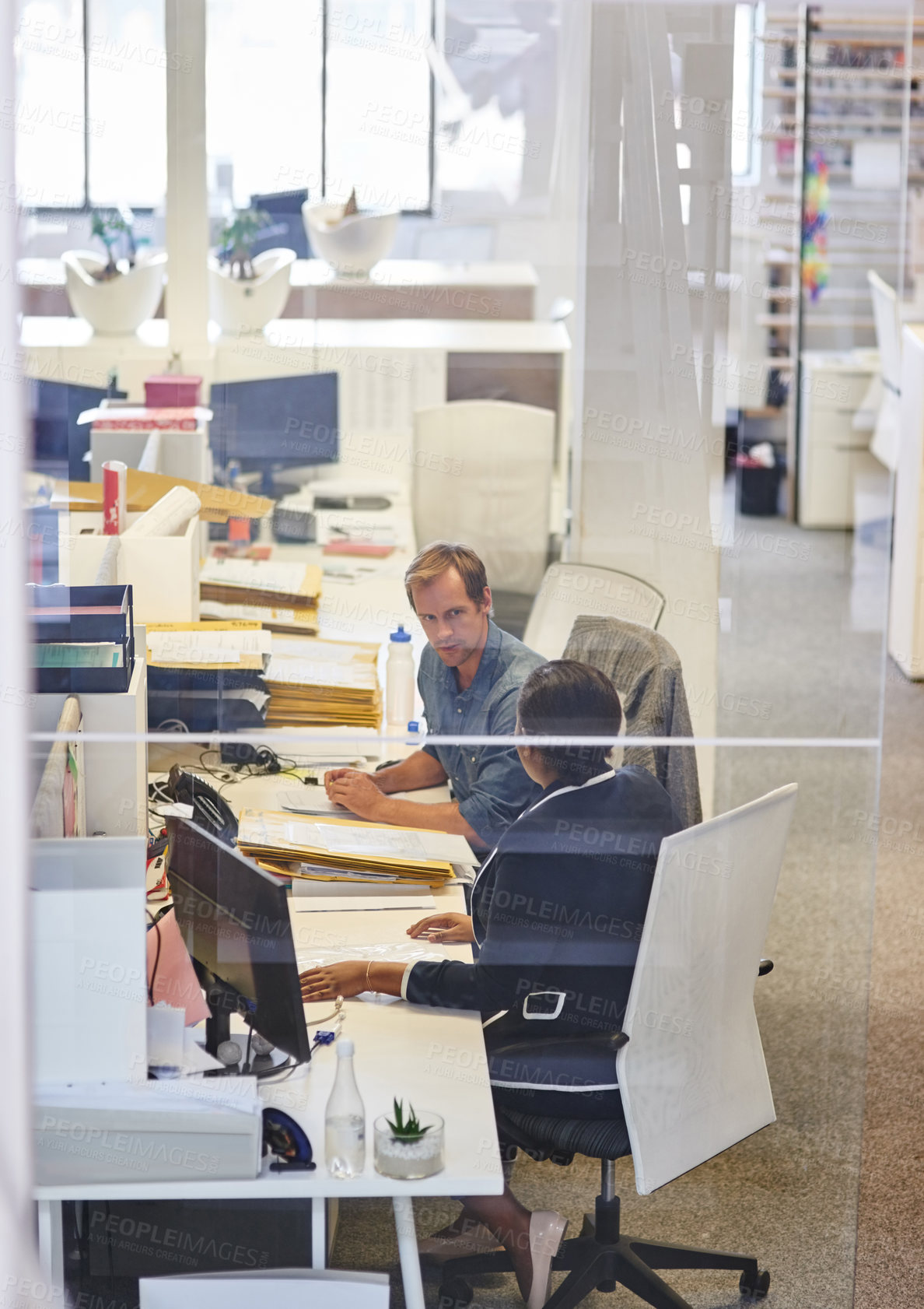 Buy stock photo Through the glass shot of two colleagues talking together in an office