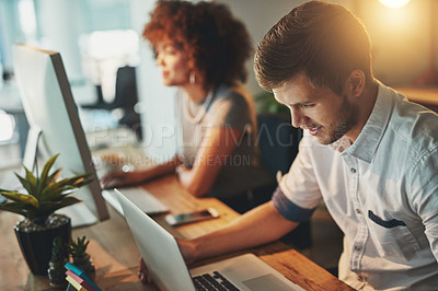 Buy stock photo Shot of a young designer working late with his colleague blurred in the background