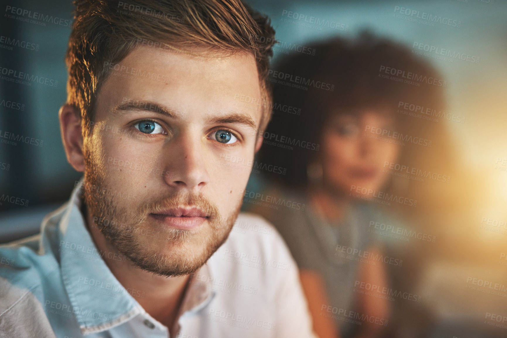 Buy stock photo Shot of a young designer working late with his colleague blurred in the background