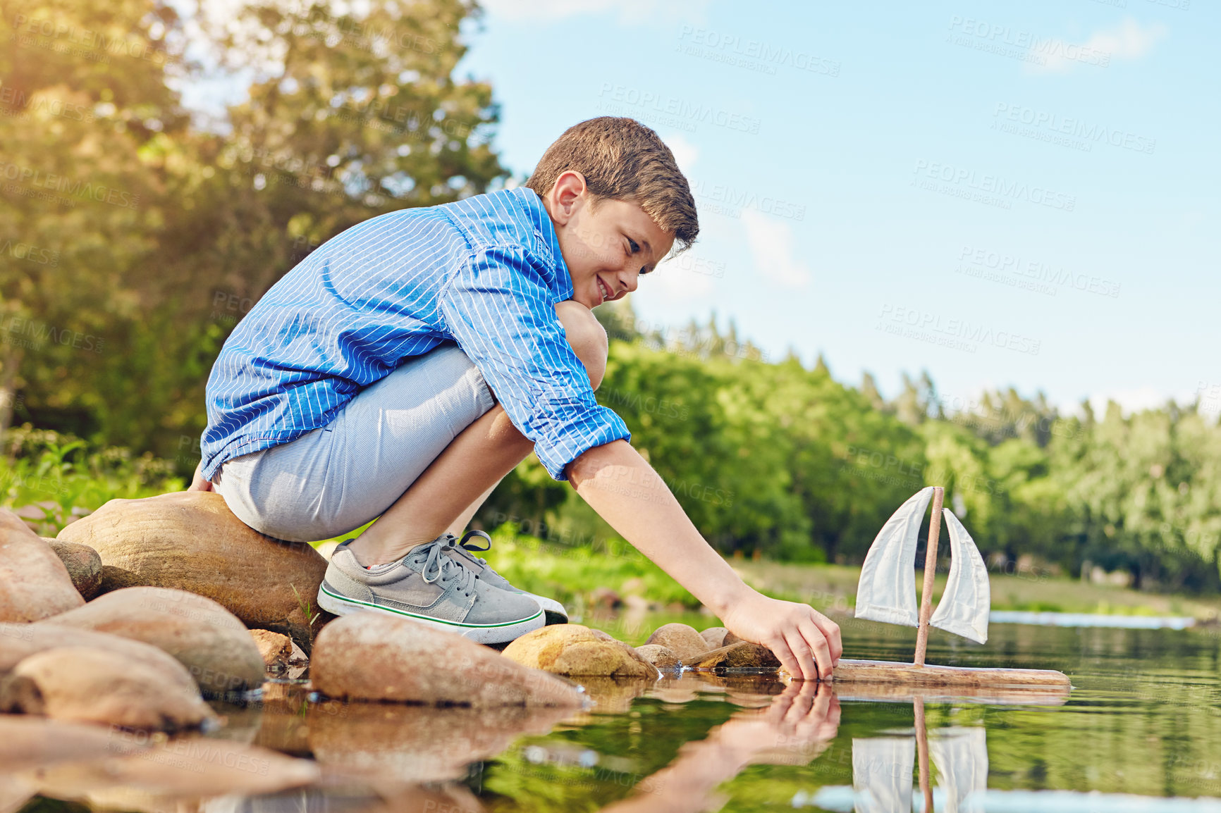 Buy stock photo Shot of a young boy playing with a toy boat by the water