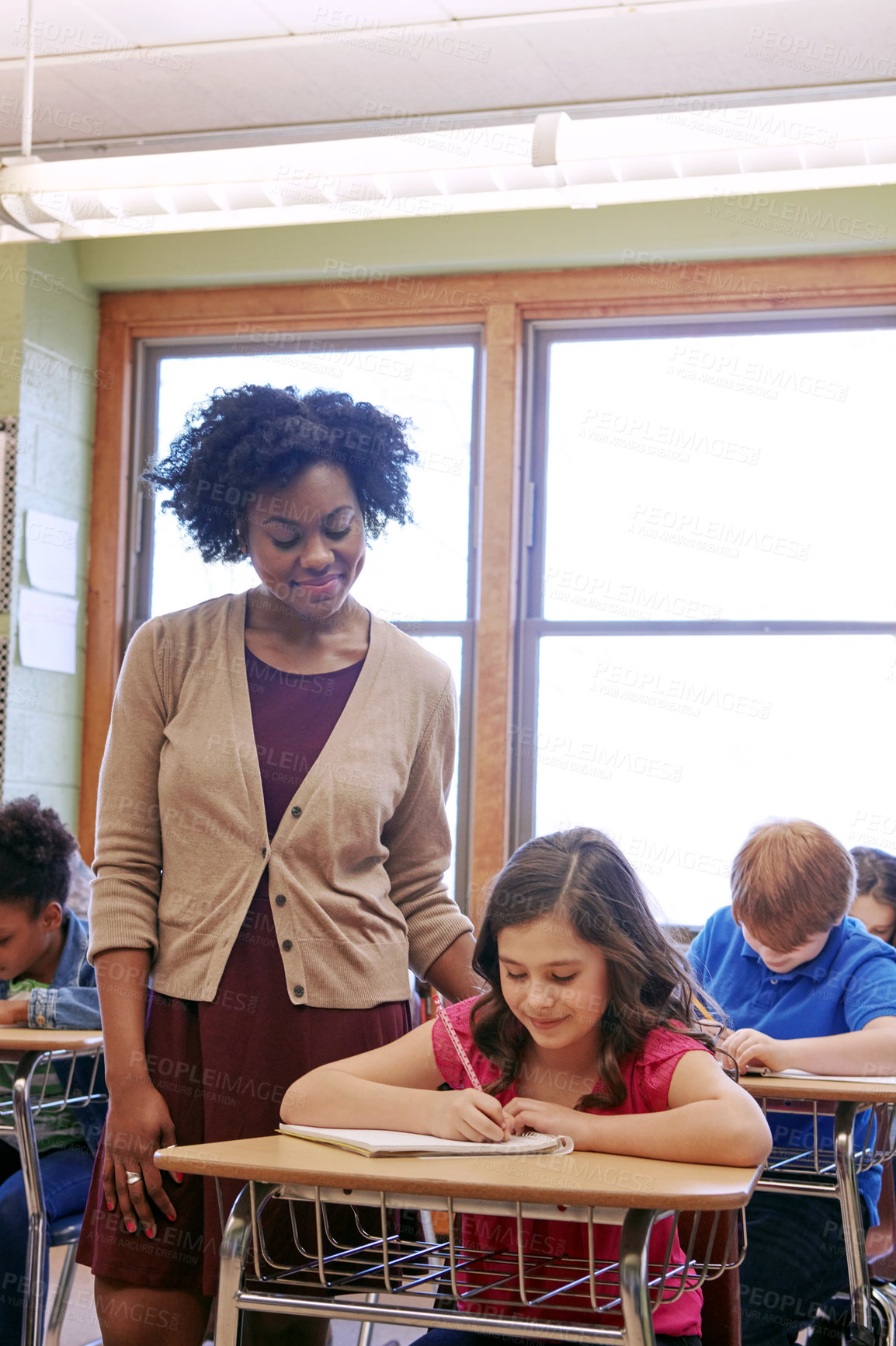 Buy stock photo Education, classroom and teacher with girl writing exam or help with test in notebook at Montessori school. Black woman, happy child at desk and monitoring students growth and development for kids.