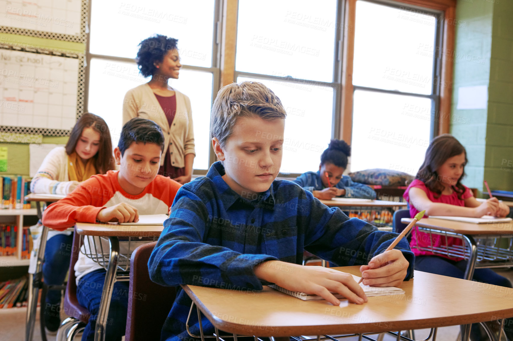 Buy stock photo Boy child, classroom and writing at desk, school and focus on education, development and learning for future. School kids, teacher and studying at table in class with diversity, notebook and pencil