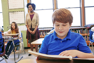 Buy stock photo Shot of a young boy sitting in class with his teacher and classmates blurred in the background