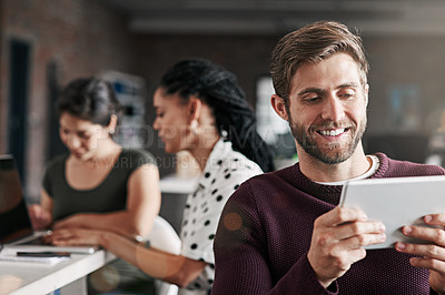 Buy stock photo Shot of a young man using a digital tablet in a modern office with his colleagues in the background