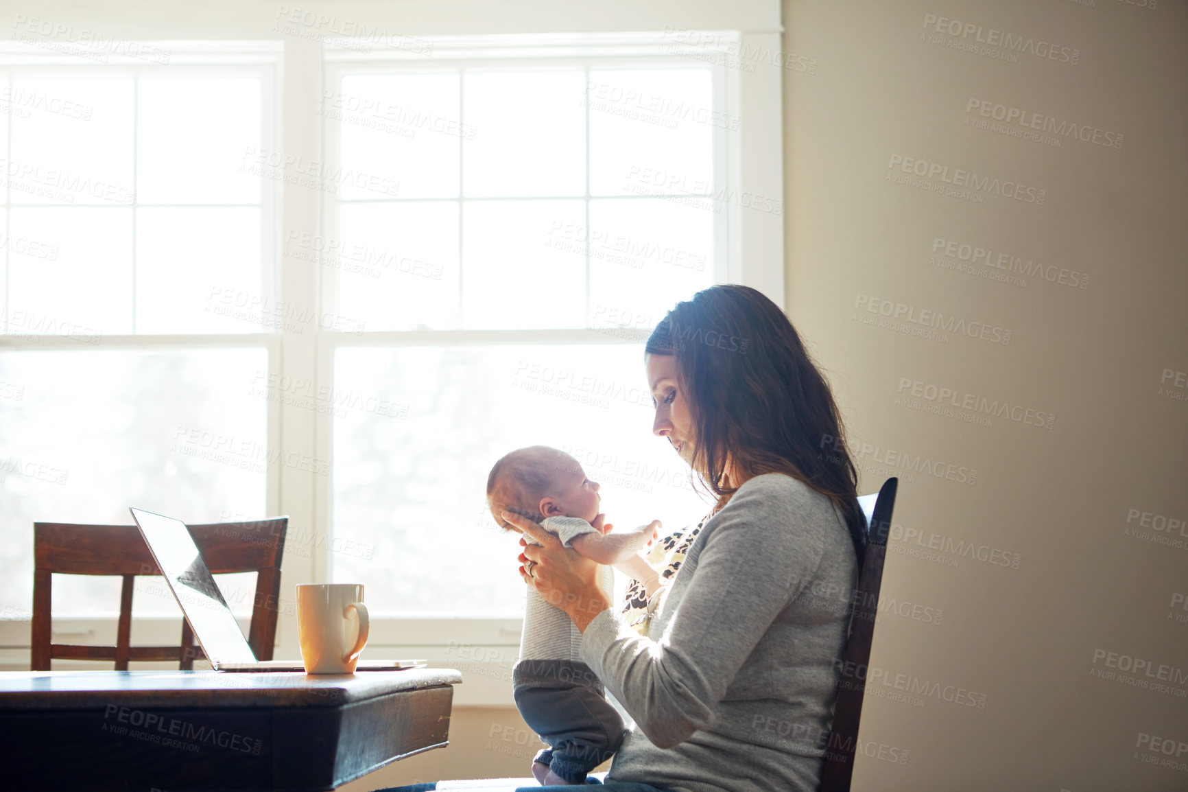 Buy stock photo Shot of a mother holding her newborn baby while sitting at a table in front of a laptop