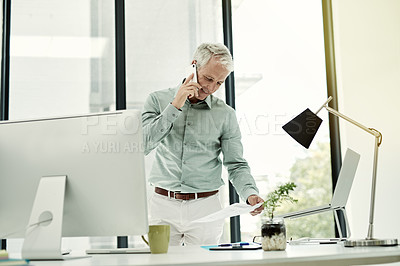 Buy stock photo Shot of a mature businessman talking on the phone while working standing at his desk in an office