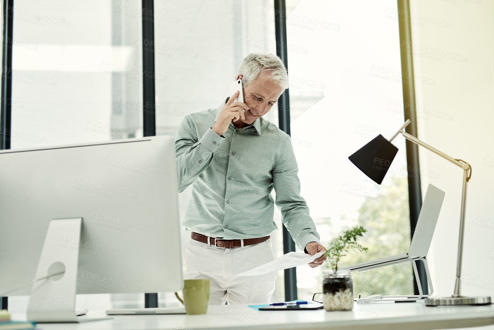 Buy stock photo Shot of a mature businessman talking on the phone while working standing at his desk in an office