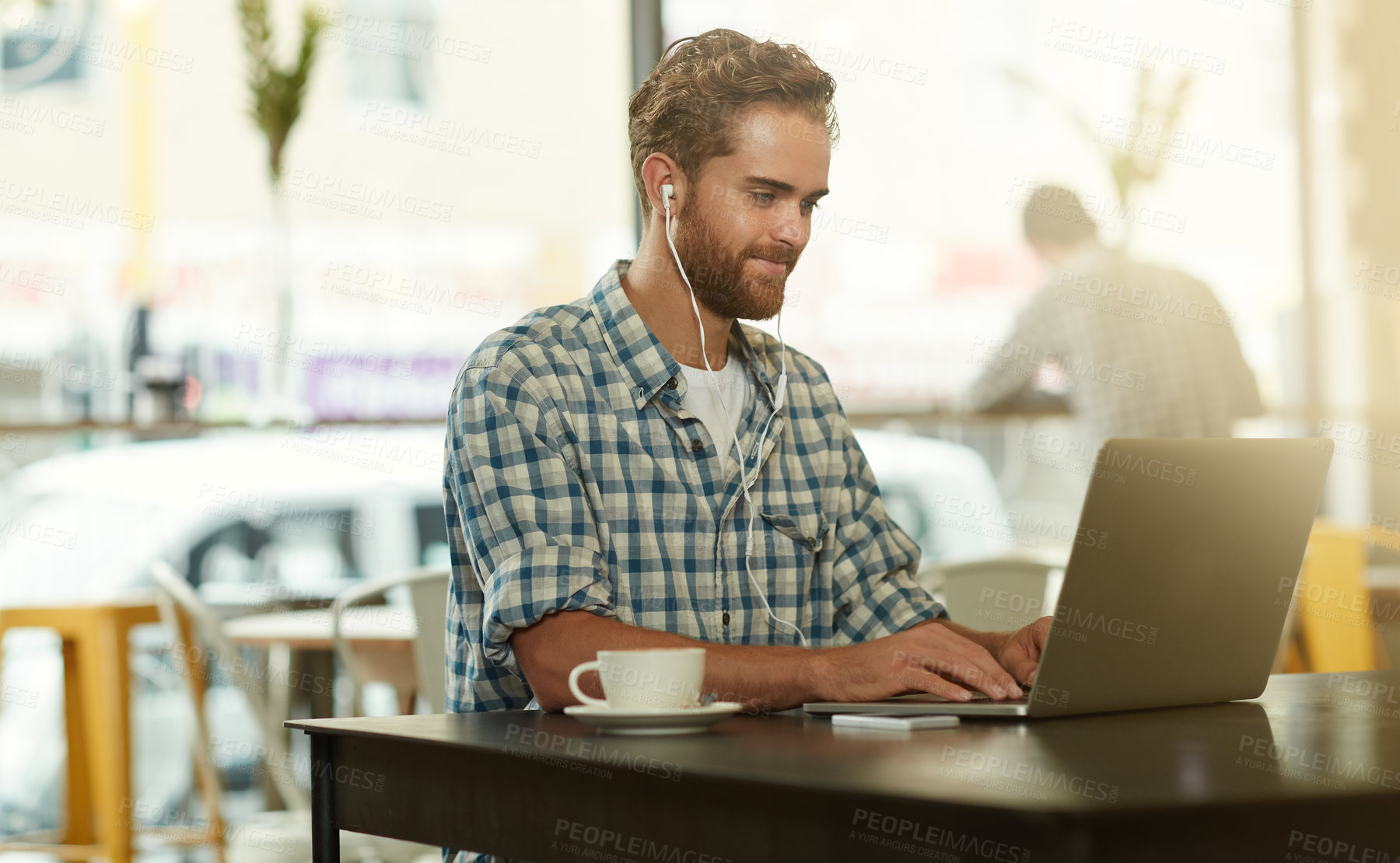 Buy stock photo Shot of a young man with earphones using a laptop in a cafe