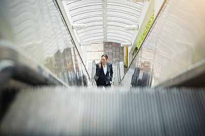 Buy stock photo Shot of a young businesswoman talking on her cellphone while standing on an escalator