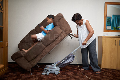 Buy stock photo Shot of a grandmother vacuuming under a couch which her grandson is lying on